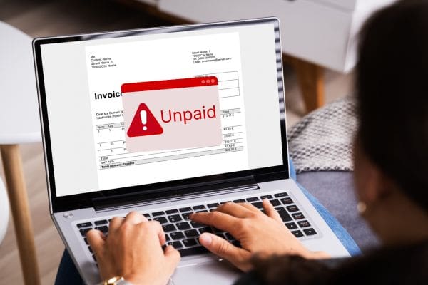 Person viewing an unpaid invoice notification on a laptop screen, with a red warning symbol indicating the invoice is overdue.